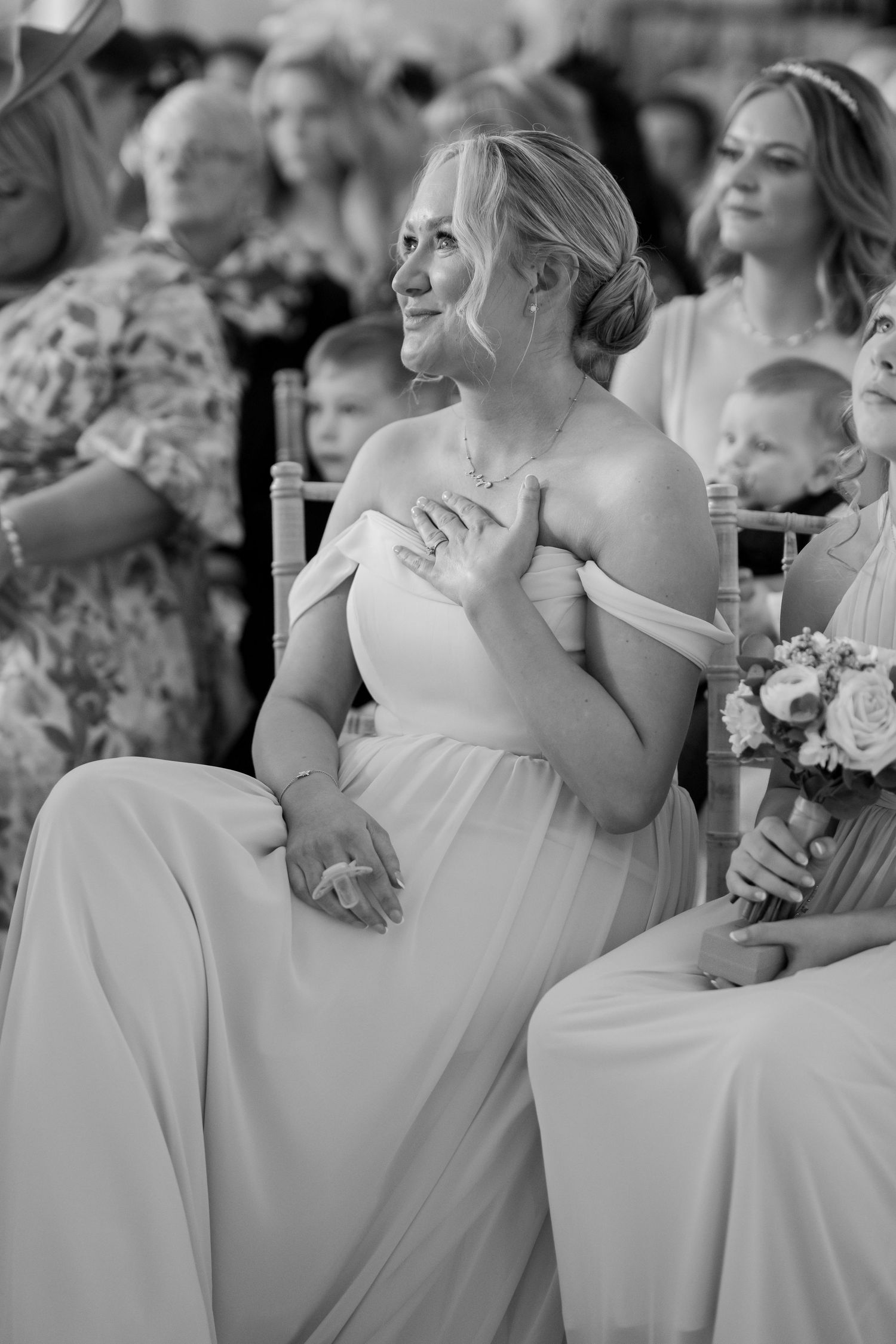 Black and white photo of an emotional bridesmaid in an off-shoulder gown, hand on chest, smiling during a wedding ceremony.