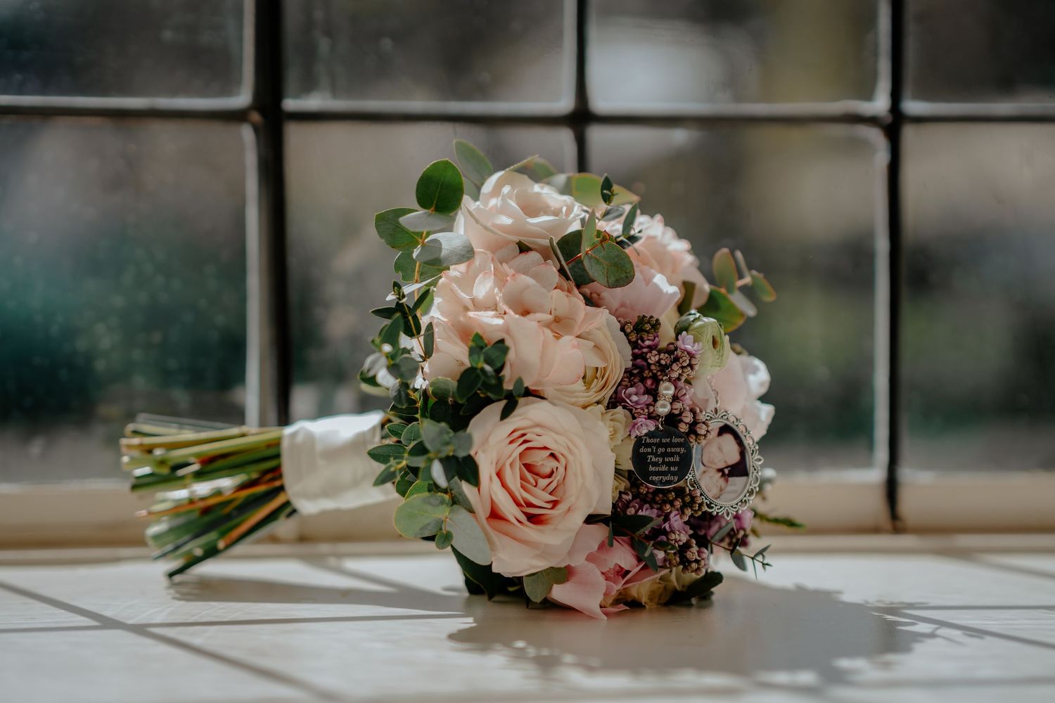 Elegant bridal bouquet of blush pink roses and greenery resting on a windowsill in soft natural light.