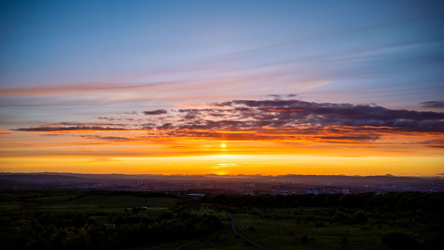 Vibrant sunset over green landscape with orange and blue sky and scattered clouds on the horizon.