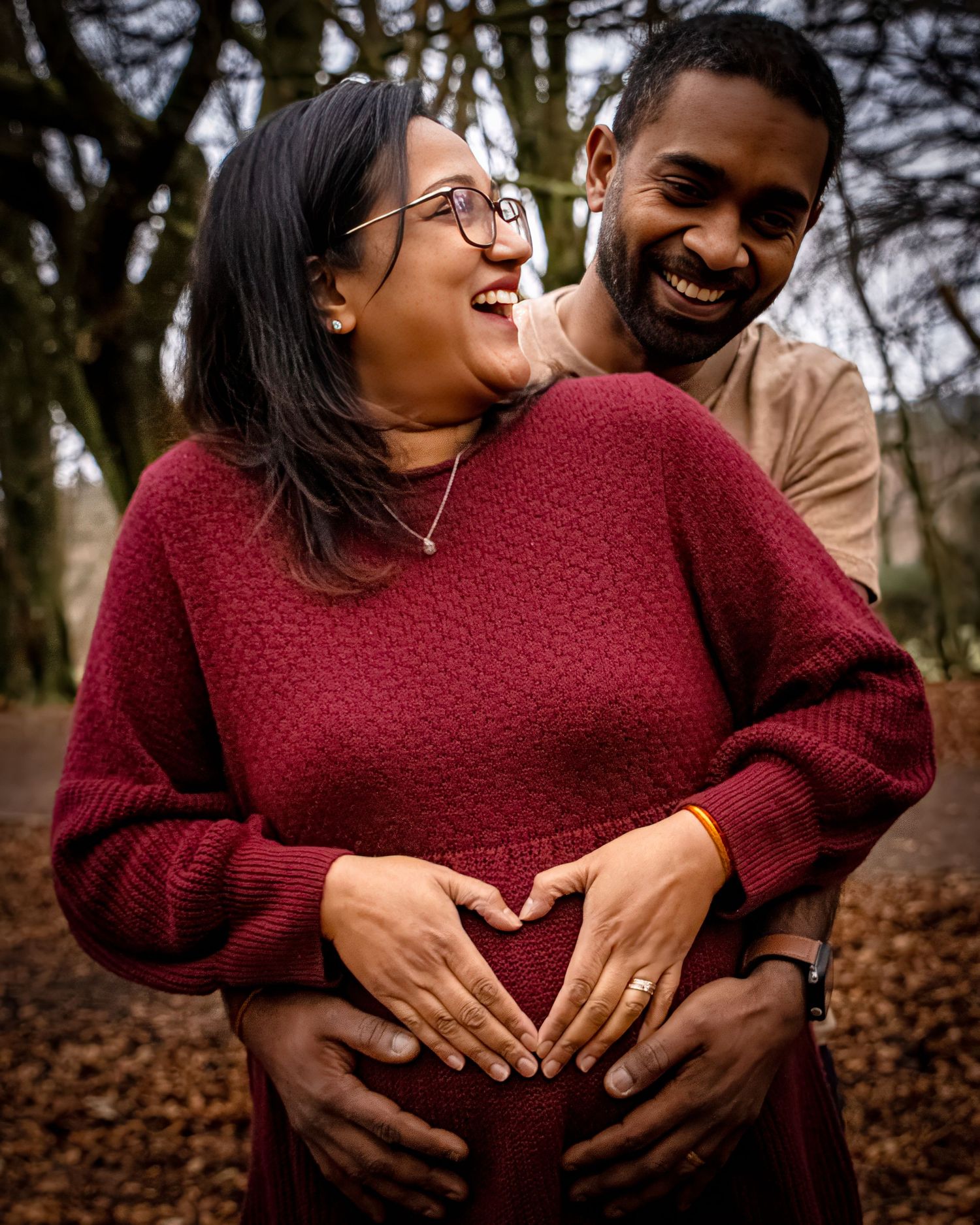 A couple shares a tender embrace while wearing burgundy and brown sweaters in an autumn setting with fallen leaves.