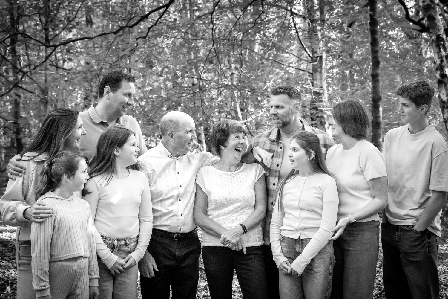 Black and white family portrait of multigenerational group laughing together outdoors in a woodland setting.