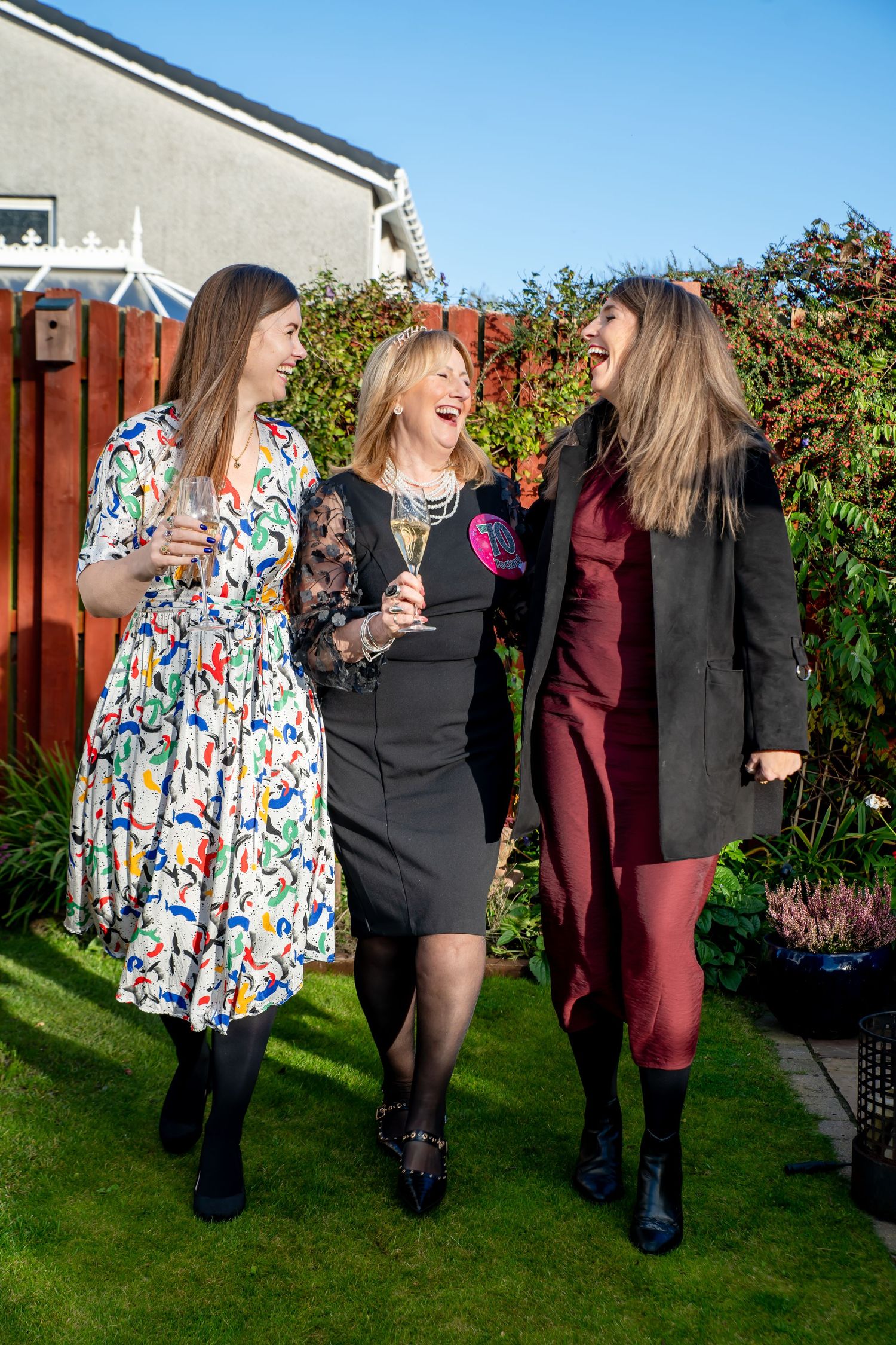 Three women laughing and walking in a garden, dressed in elegant outfits, holding champagne glasses on a sunny day.