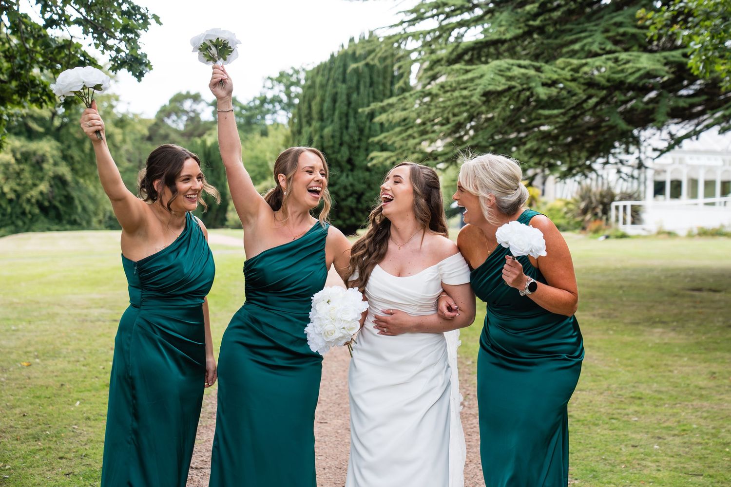 Bride in white gown laughing with three bridesmaids in emerald green dresses, celebrating outdoors on a sunny wedding day.
