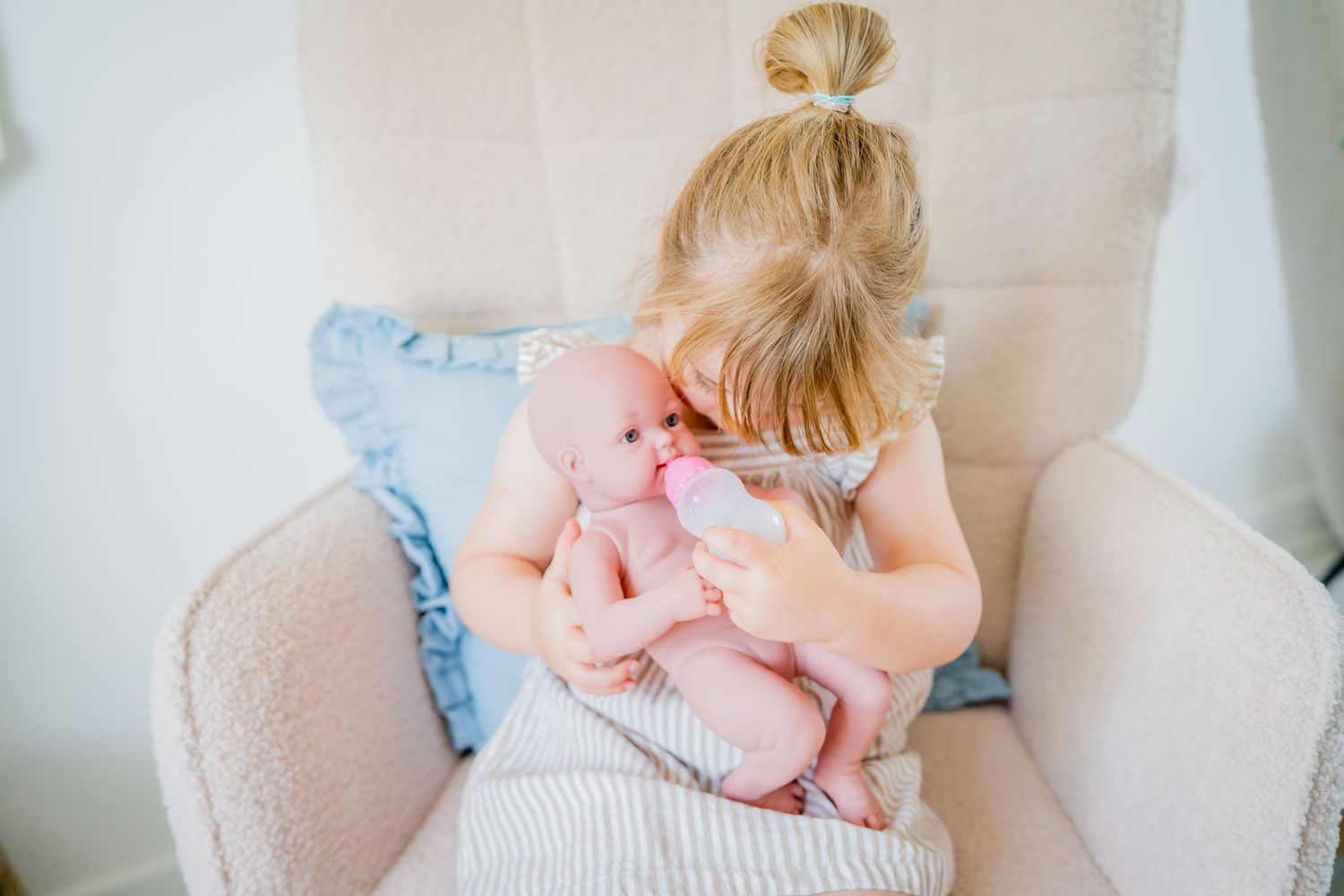 A young blonde girl sits in a beige armchair, lovingly cradling and kissing a baby doll.