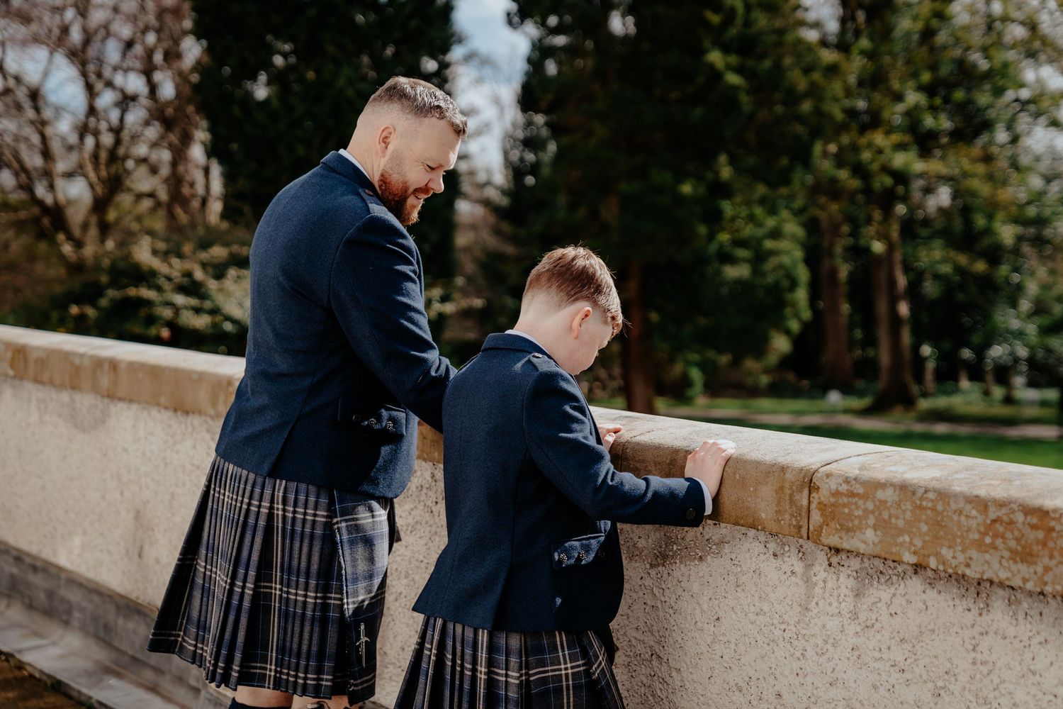 Two males in matching navy kilts and blazers stand at a stone balustrade in a scenic garden setting.