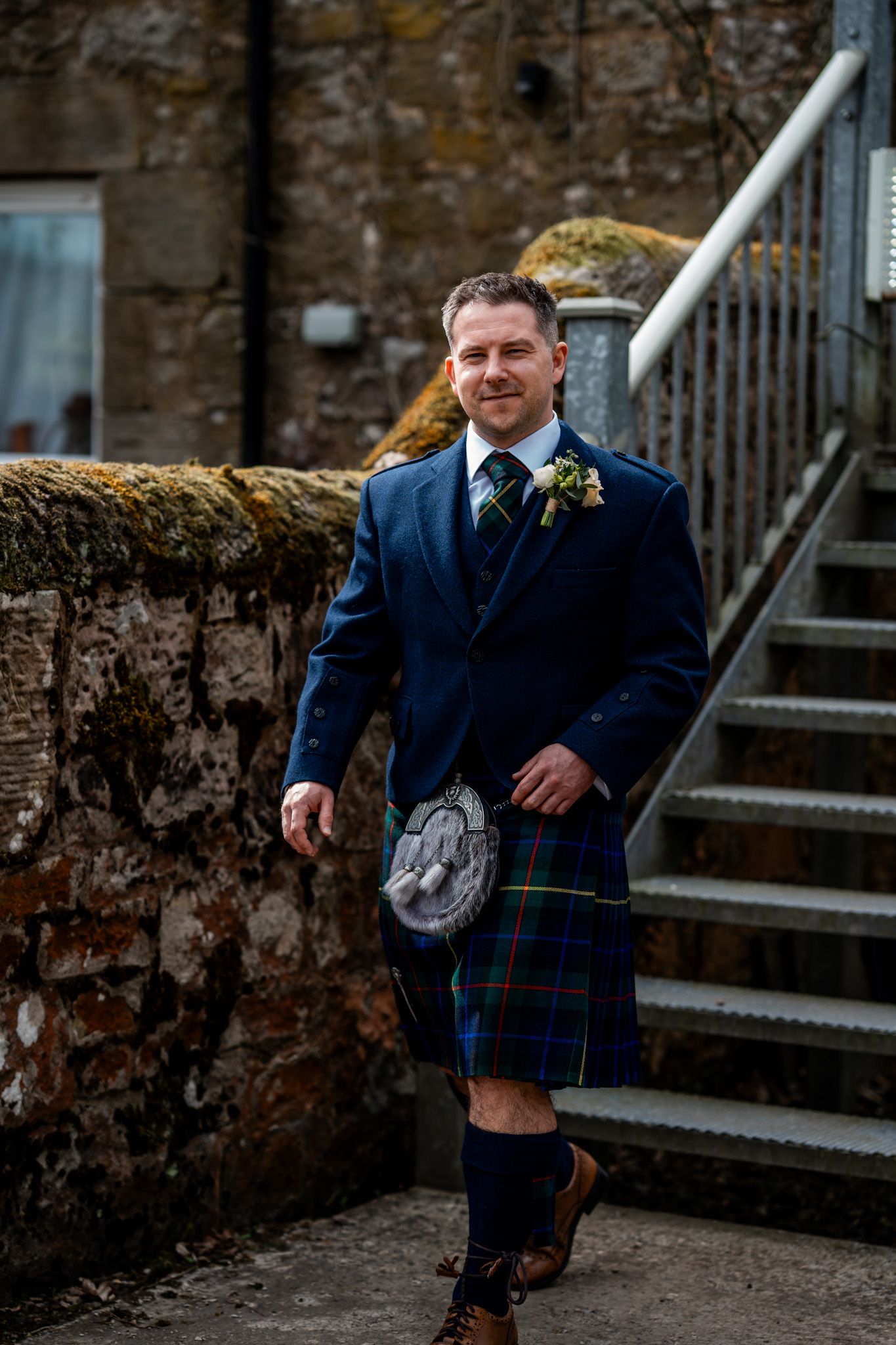 Groom in blue jacket and tartan kilt with sporran and boutonniere stands by stone steps at Scottish wedding.