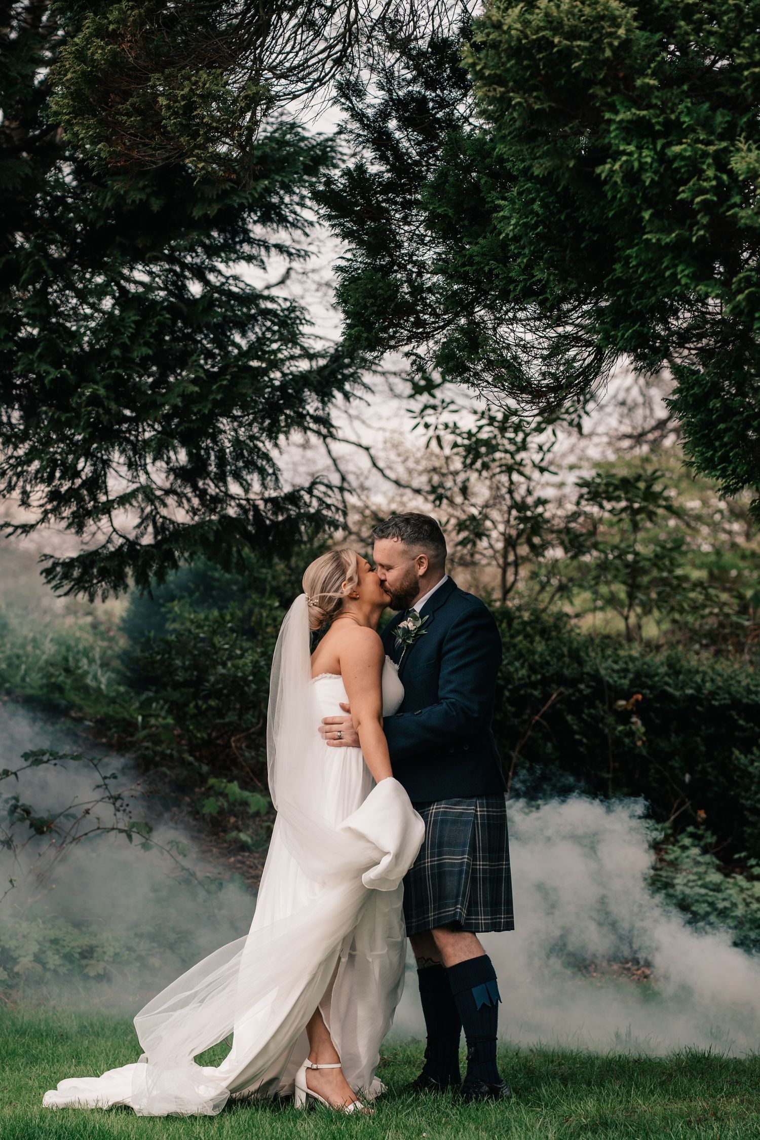Bride and groom sharing a romantic kiss under lush trees, groom wearing a traditional Scottish kilt, surrounded by soft mist.