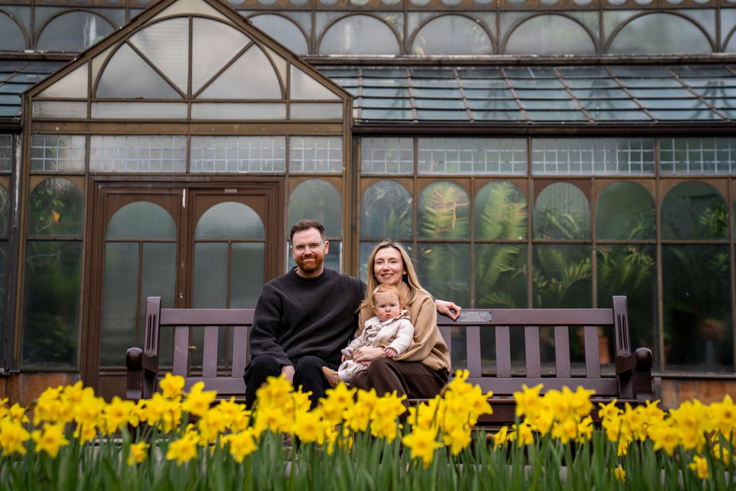 Couple sitting on a wooden bench with a baby, surrounded by yellow daffodils in front of a glass greenhouse.