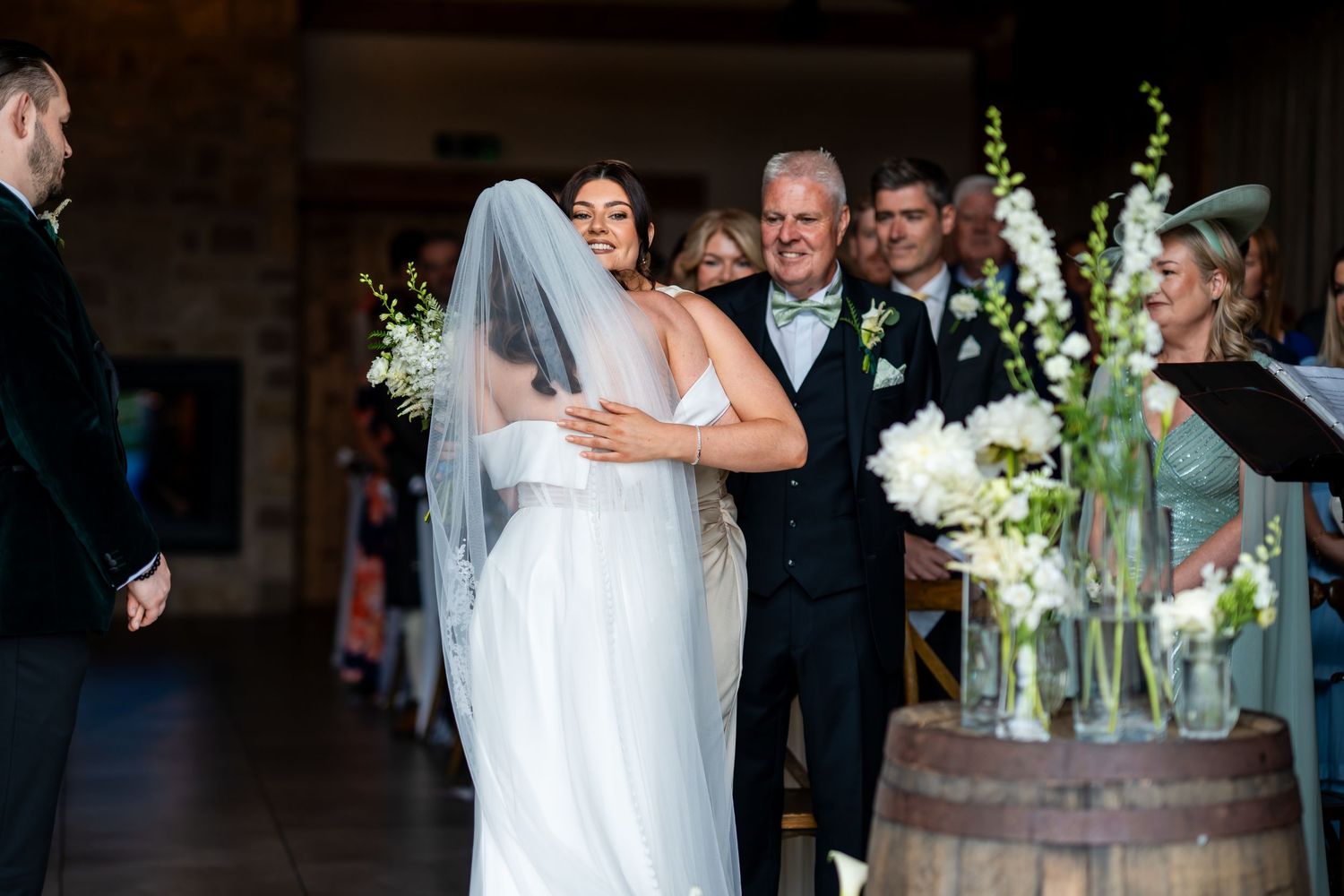 Bride walking down the aisle with her father during an elegant indoor wedding ceremony with white floral arrangements.