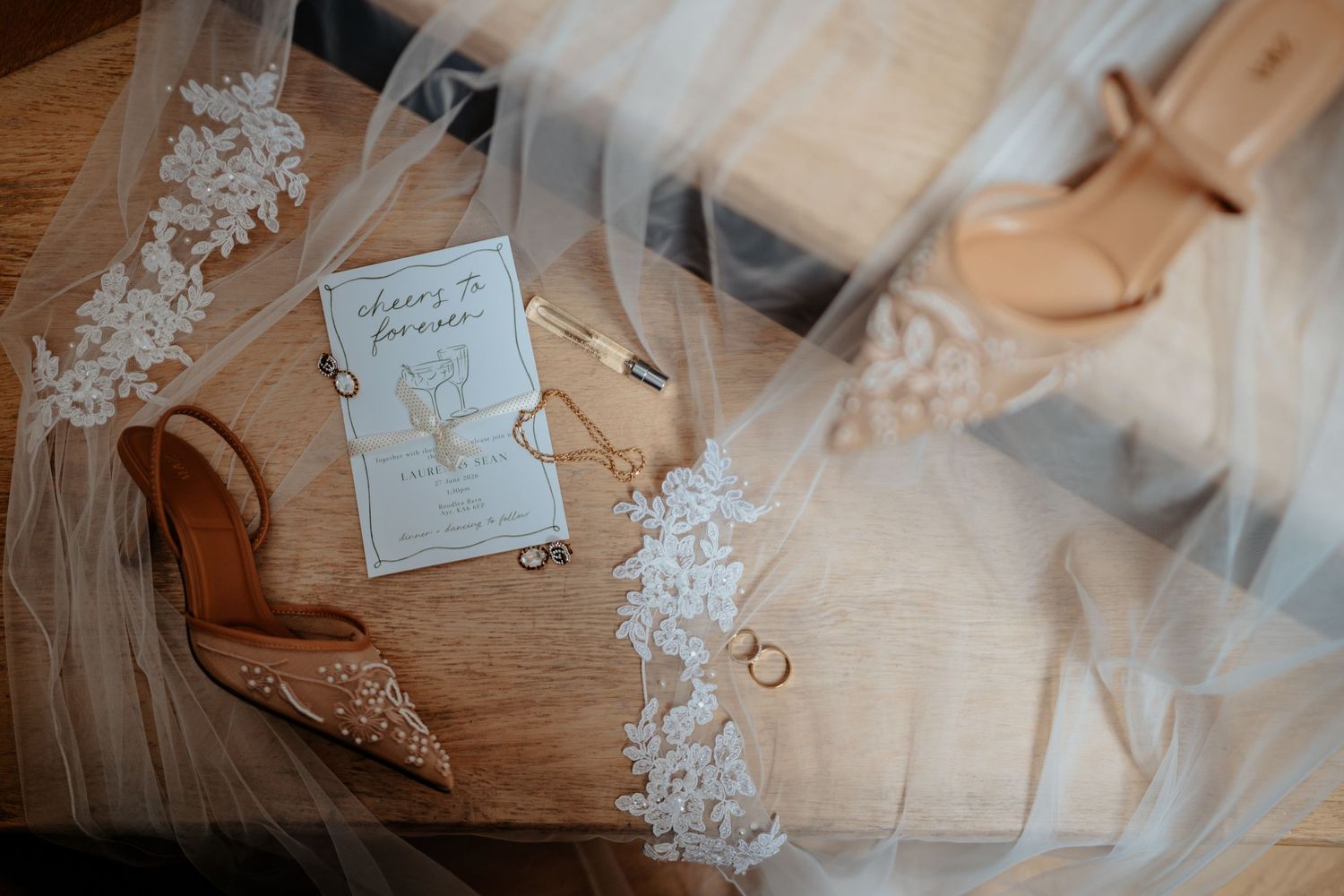 Bridal flat lay with lace veil, nude heels, gold rings, and wedding invitation on a wooden surface.
