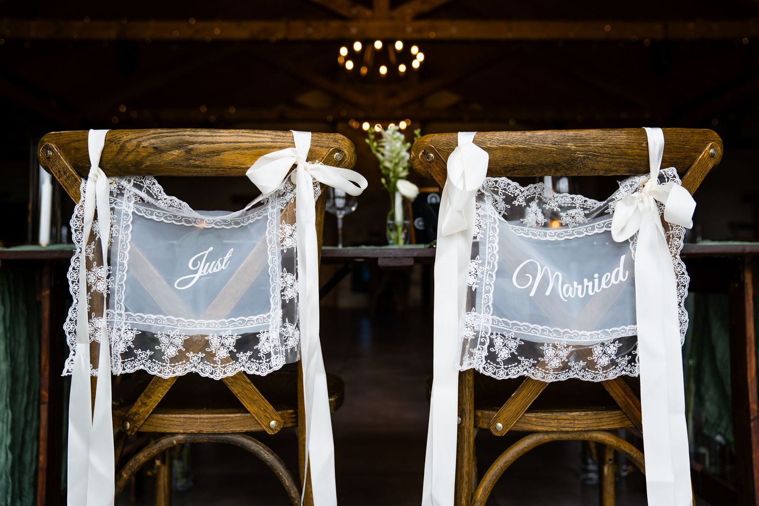 Just Married signs on rustic wooden wedding chairs decorated with white lace and ribbon bows at a barn venue.