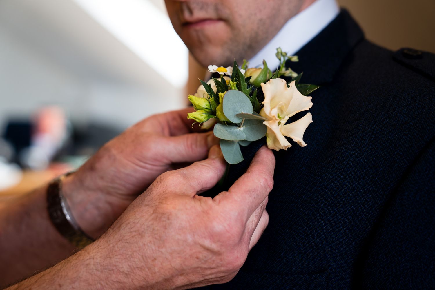 Hands pinning a white floral boutonniere with eucalyptus onto a groom's dark suit lapel.