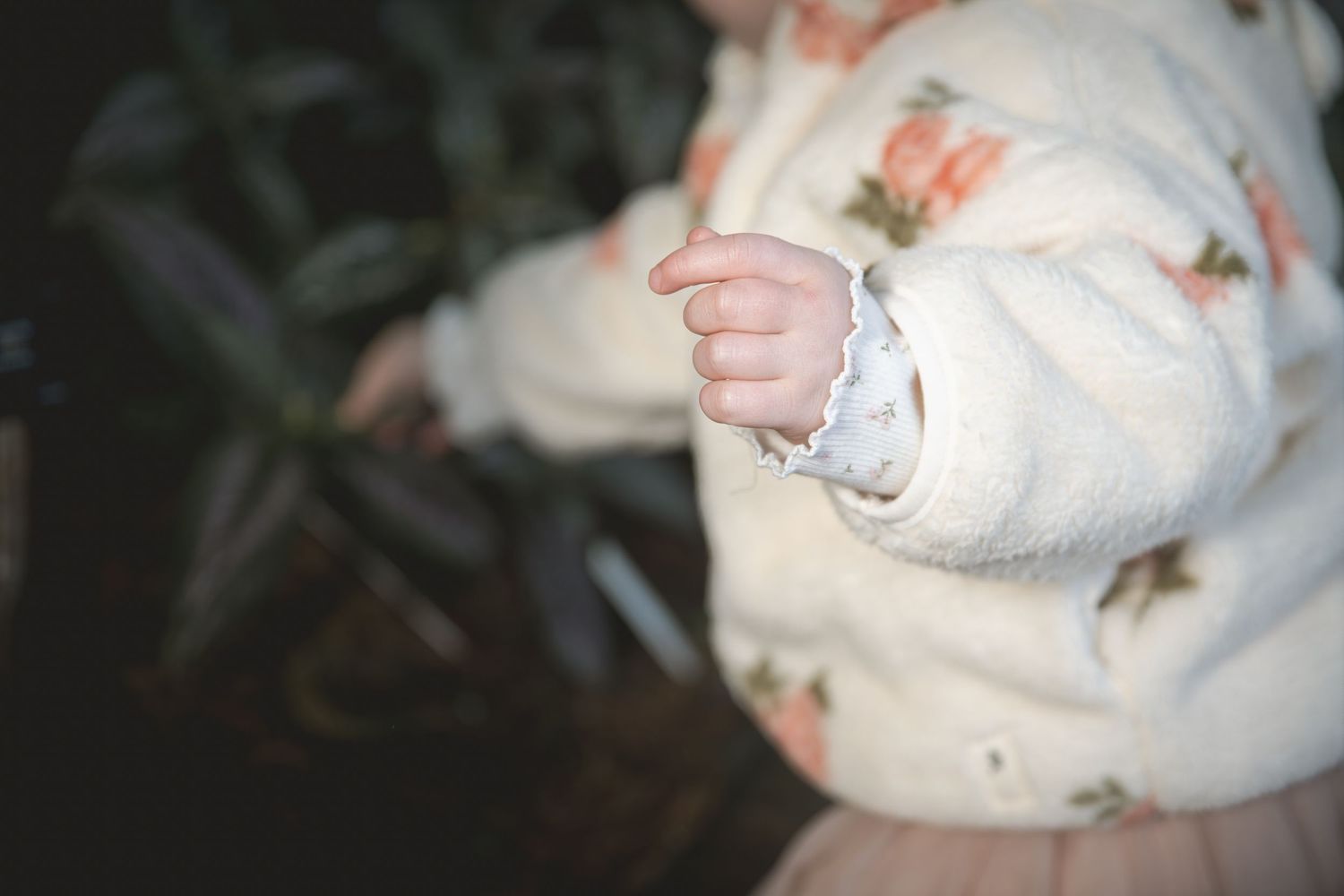 Close-up of a baby's hand in a floral white fuzzy jacket with a lace cuff, outdoors near green plants.