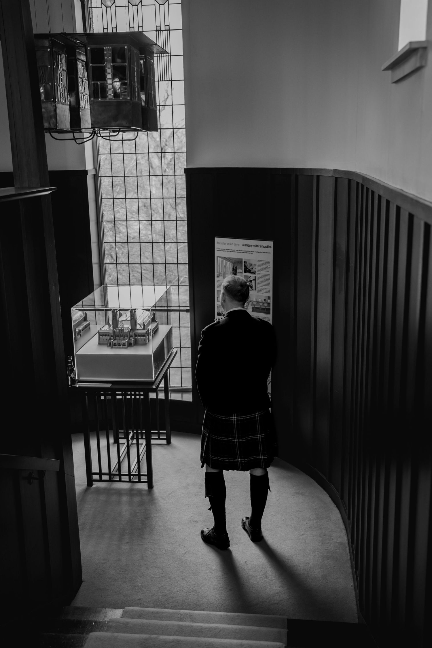 Black and white photo of a man in a tartan kilt standing at the top of a staircase in a historic building.