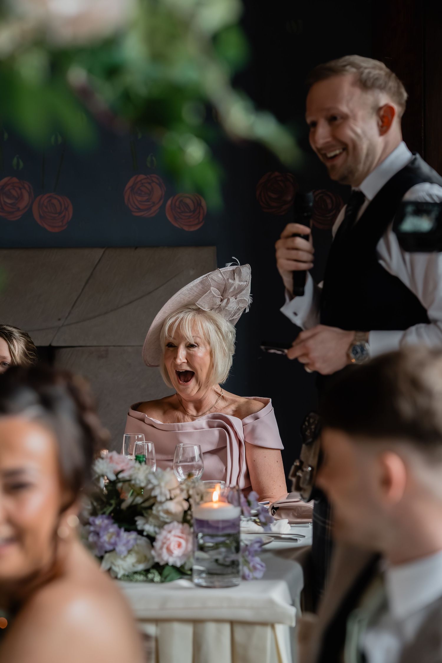 Wedding guest in fascinator hat reacts with surprise during speech at elegant reception dinner.