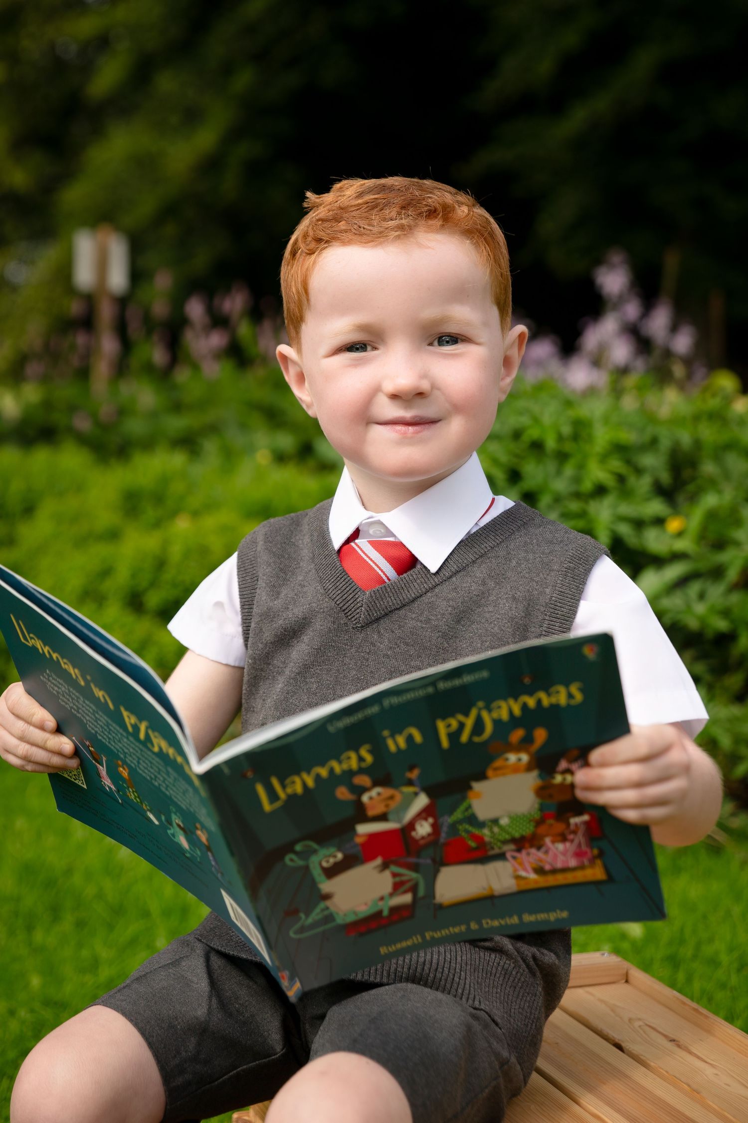 Red-haired boy in school uniform reads Llamas in Pyjamas book outdoors in a lush green garden setting.