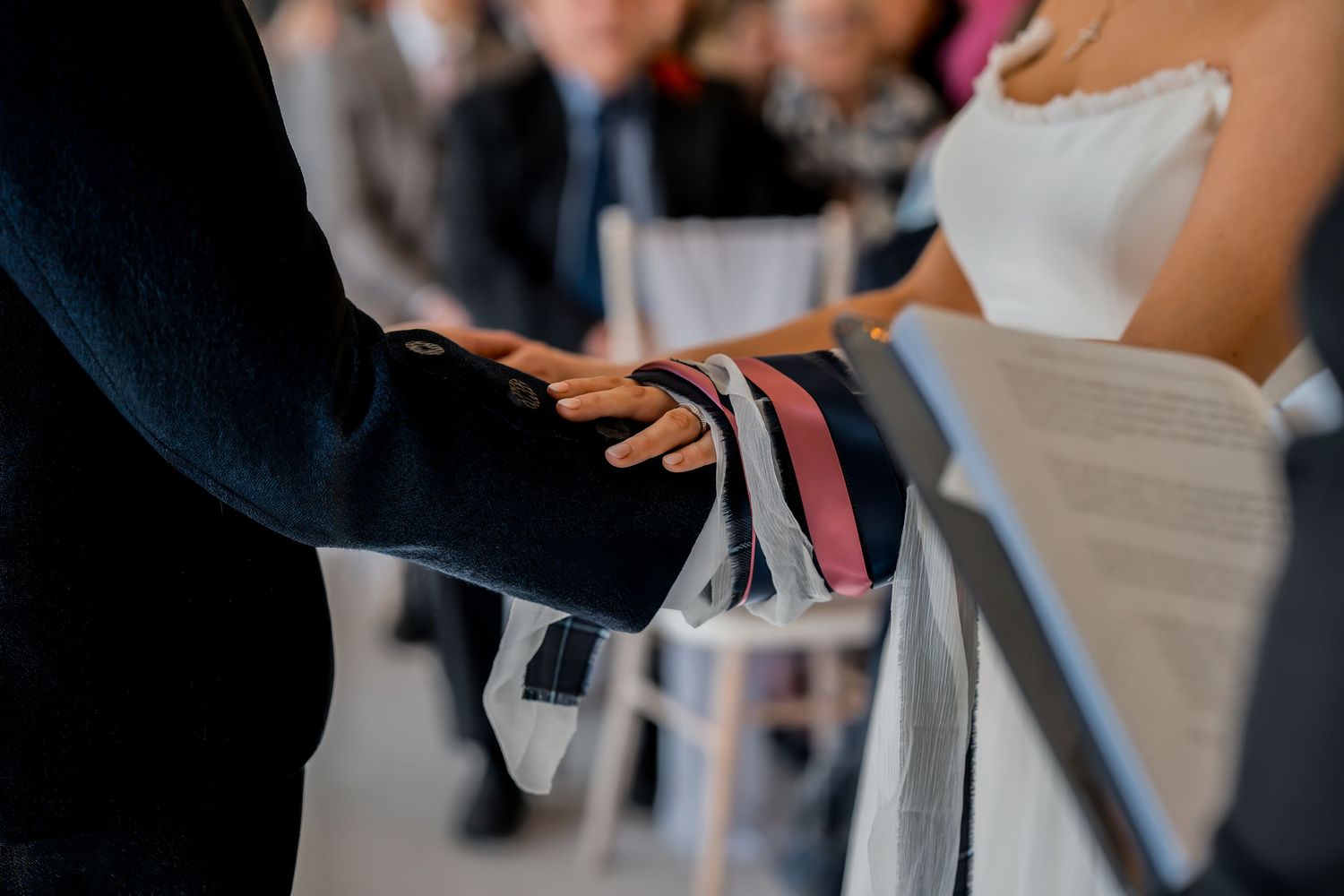 Close-up of hands exchanging wedding vows ribbons during a romantic ceremony with guests seated in background.