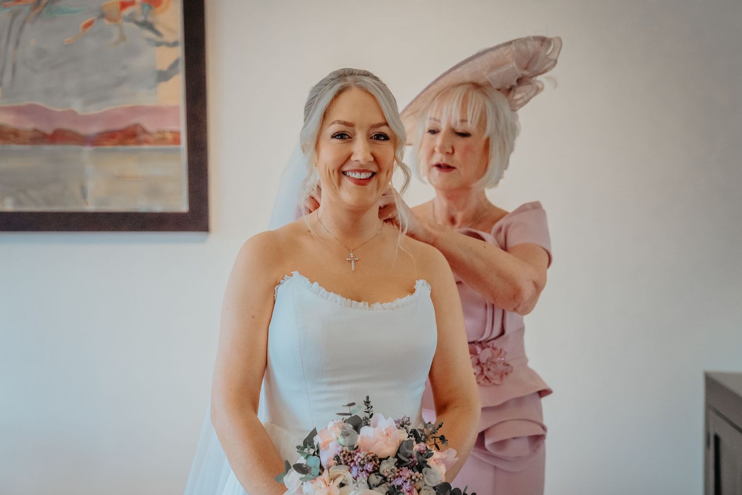 Bride holding bouquet gets necklace fastened by older woman in pink dress and fascinator during wedding preparations.