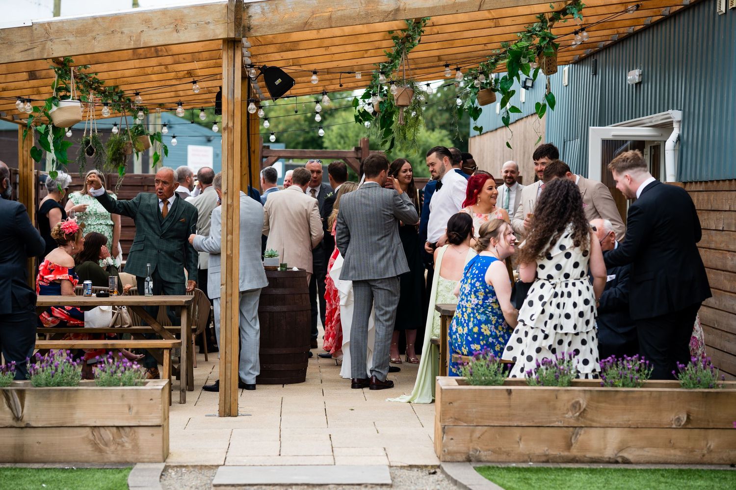 Guests mingle outdoors at a garden wedding reception under a wooden pergola adorned with greenery.