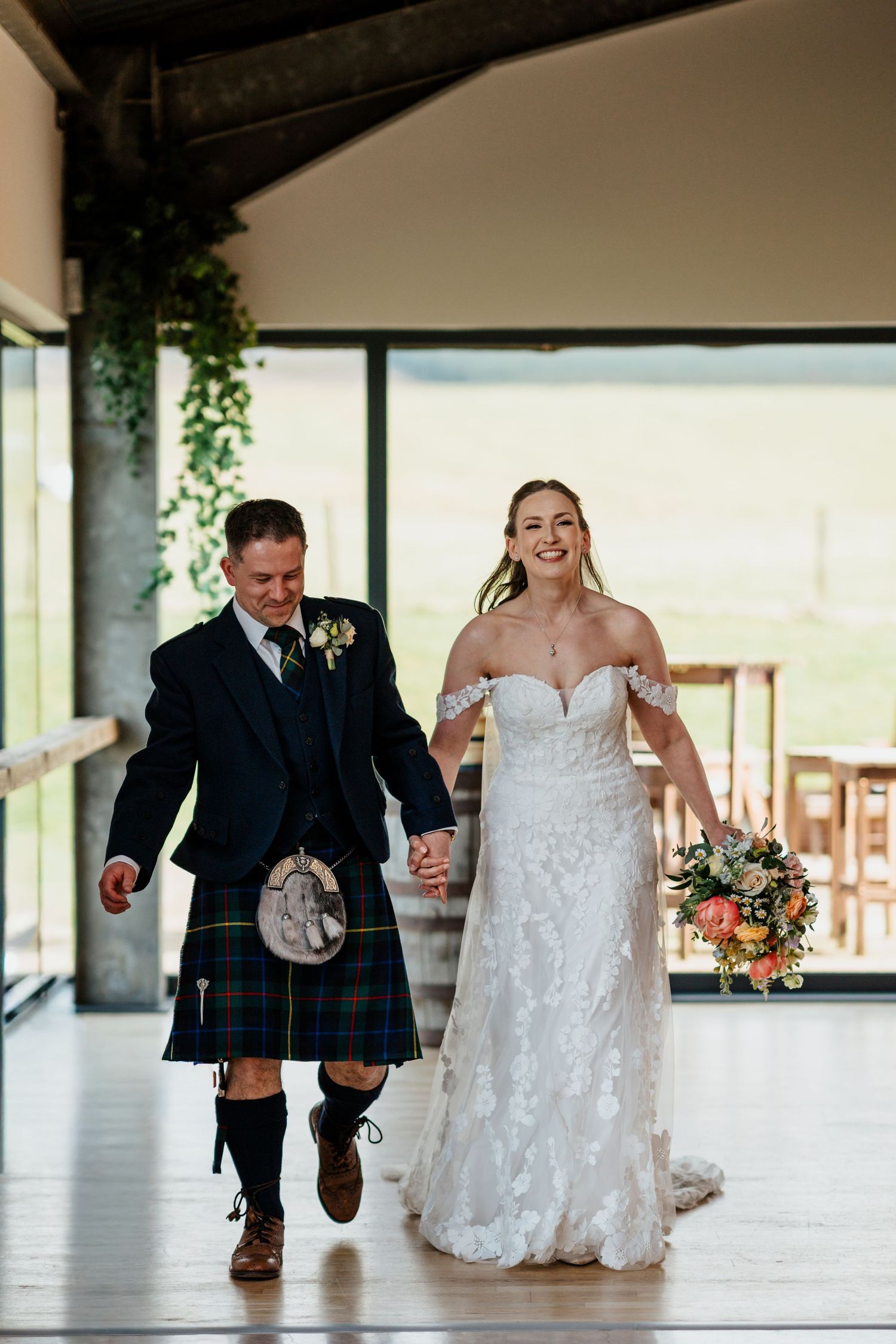 Bride in lace off-shoulder gown holds bouquet while walking with man in traditional Scottish kilt at wedding venue.