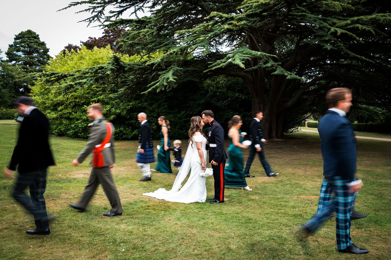 Wedding couple kissing on lawn while guests in kilts and formal wear walk through scenic garden grounds.