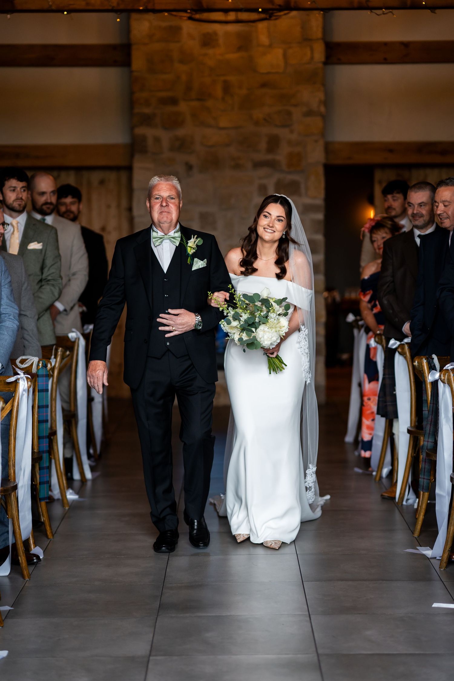 Bride walks down the aisle with her father in a rustic barn wedding venue, holding a white floral bouquet.