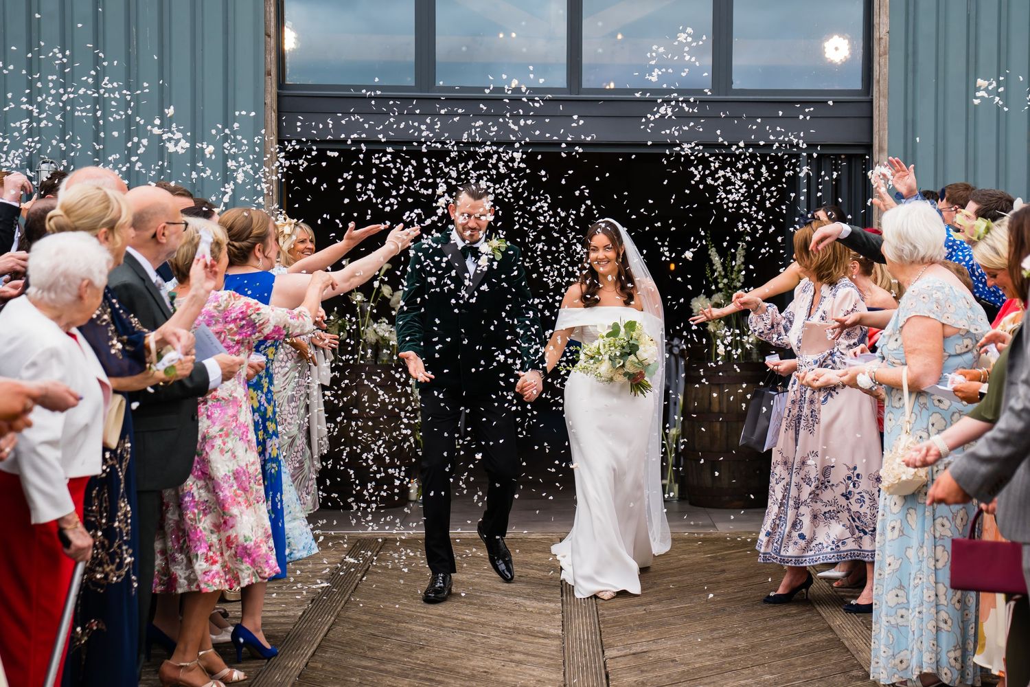Bride and groom walk through confetti shower surrounded by cheering wedding guests outside a barn venue.