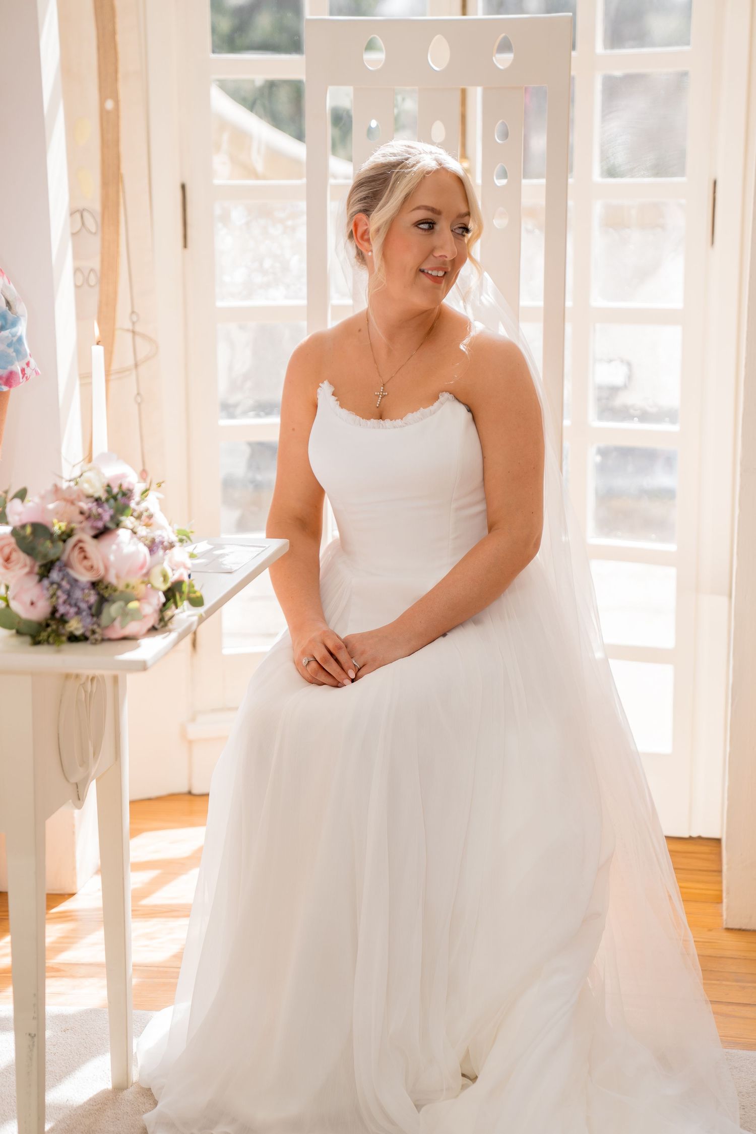 Bride in strapless white tulle wedding gown standing by bright French doors with floral bouquet nearby.