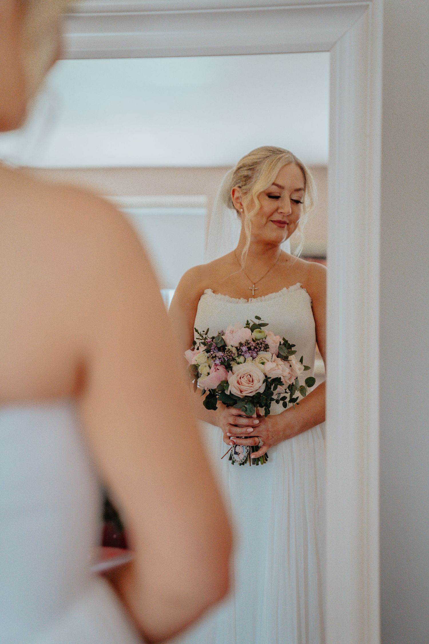 Blonde bride in strapless white gown holds pink rose bouquet while admiring her reflection in a mirror.