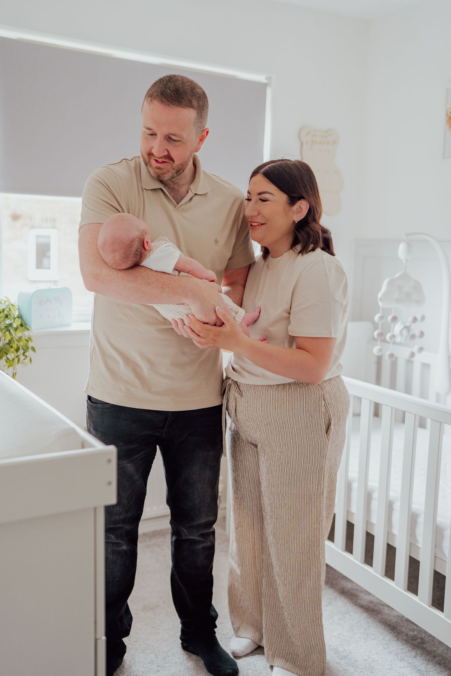 Happy couple standing in nursery holding their newborn baby near a white crib in a bright, modern room.