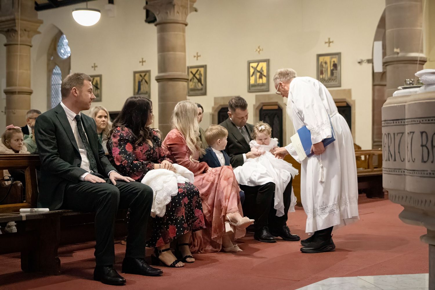 A priest performs a baptism ceremony for families seated in church pews near a stone baptismal font.