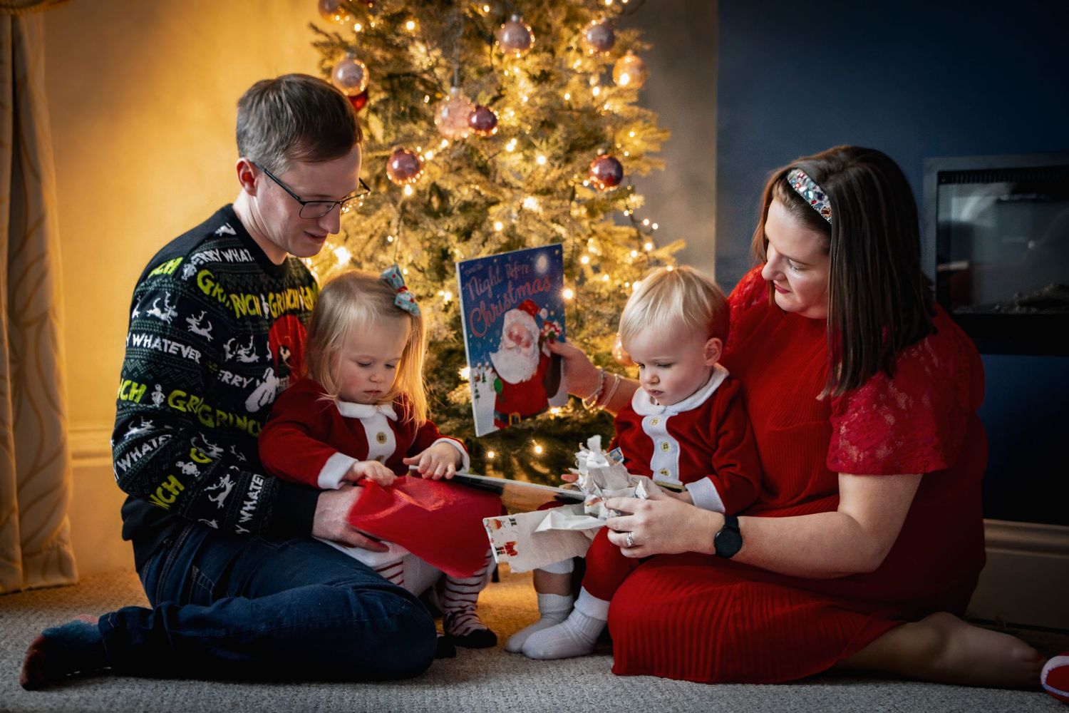 Family of four celebrating Christmas together near a decorated tree, parents and two toddlers opening gifts in festive attire.