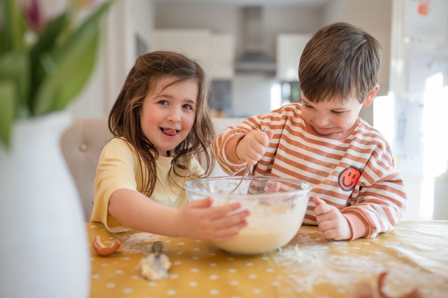 Two happy children mixing batter in a glass bowl at a kitchen table, enjoying baking together at home.