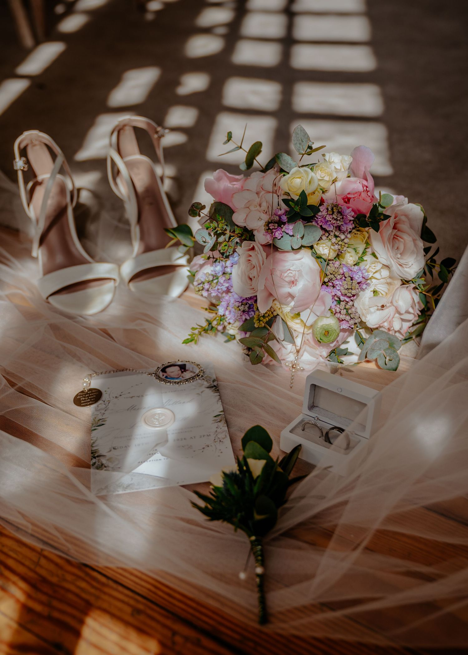 Elegant wedding flat lay with blush floral bouquet, bridal heels, boutonniere, and stationery on tulle fabric.