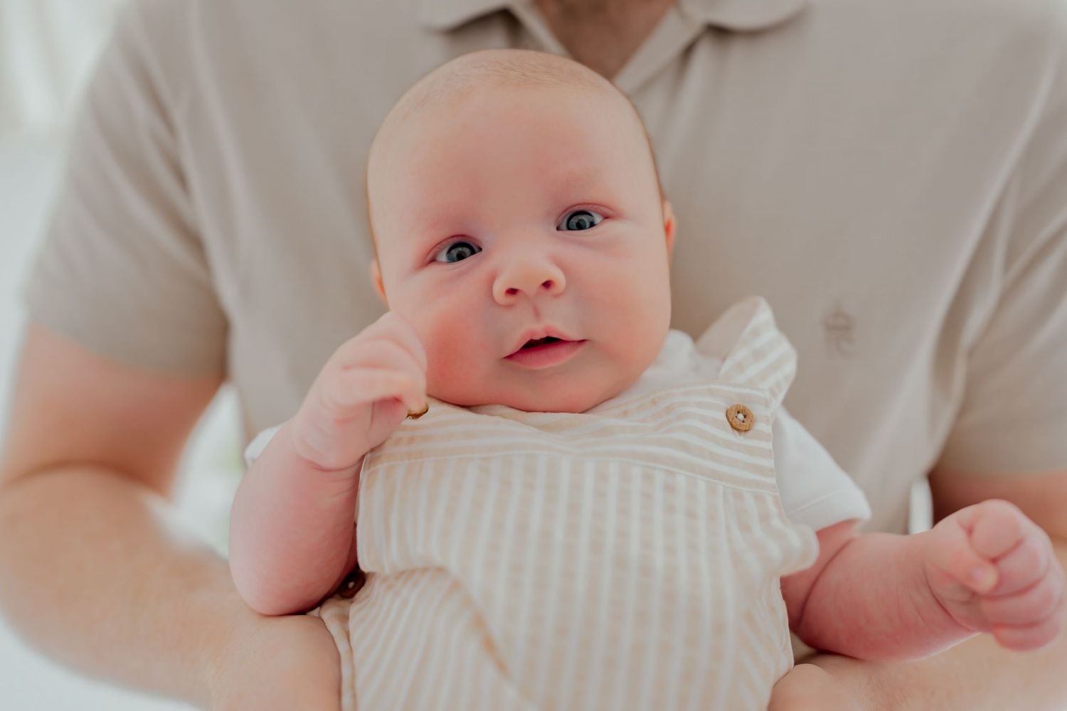 Newborn baby in white striped romper being held by father in beige shirt during professional photoshoot.