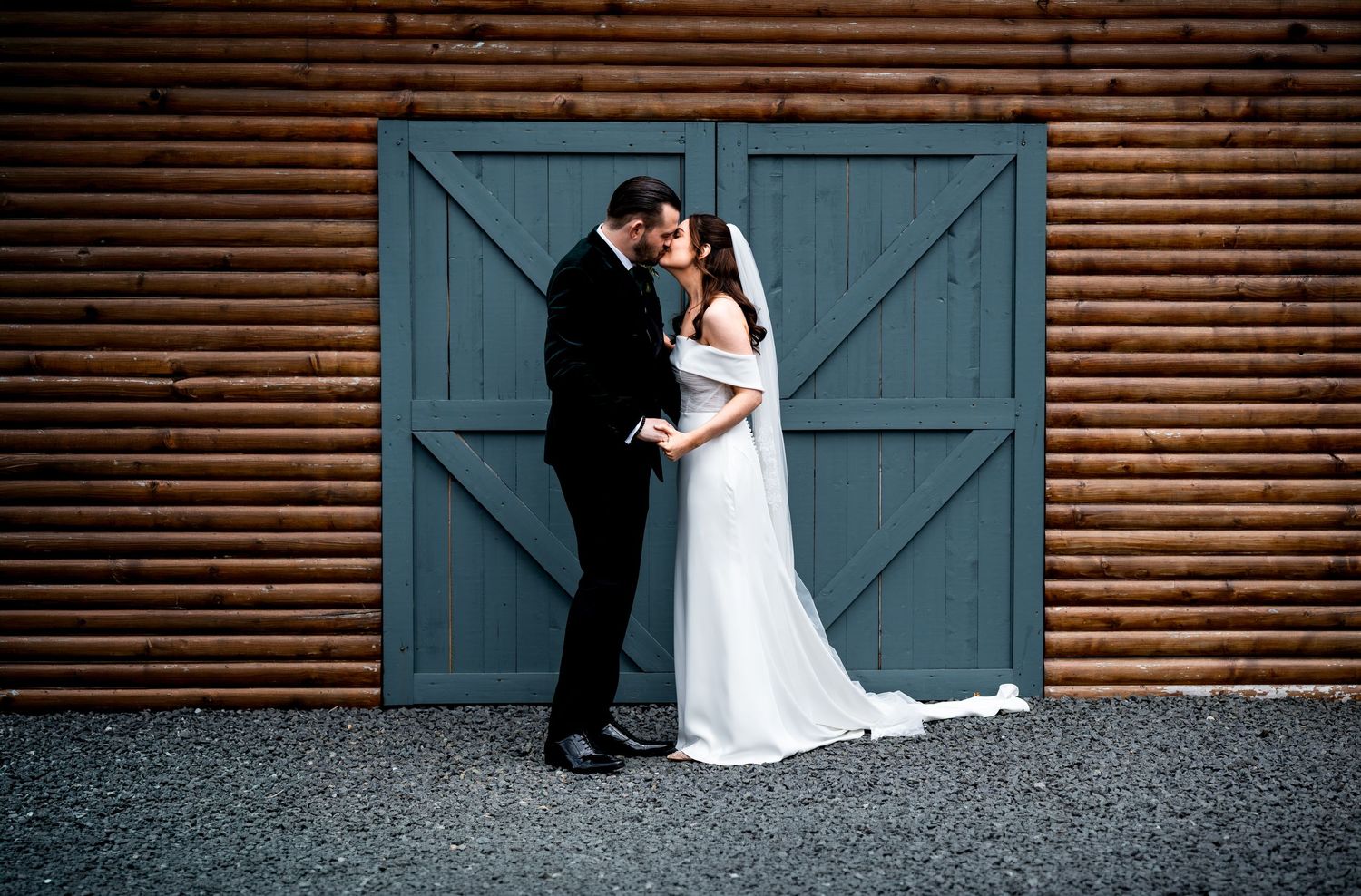 Bride and groom share a romantic kiss in front of rustic log barn with teal doors on their wedding day.