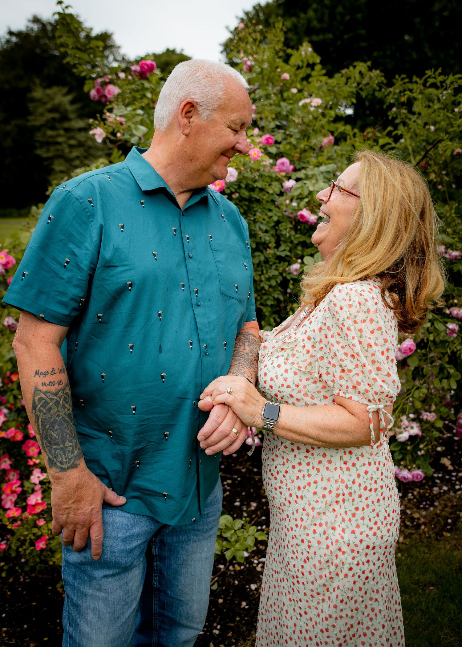 Couple in a garden with pink roses, wearing teal and floral print outfits.