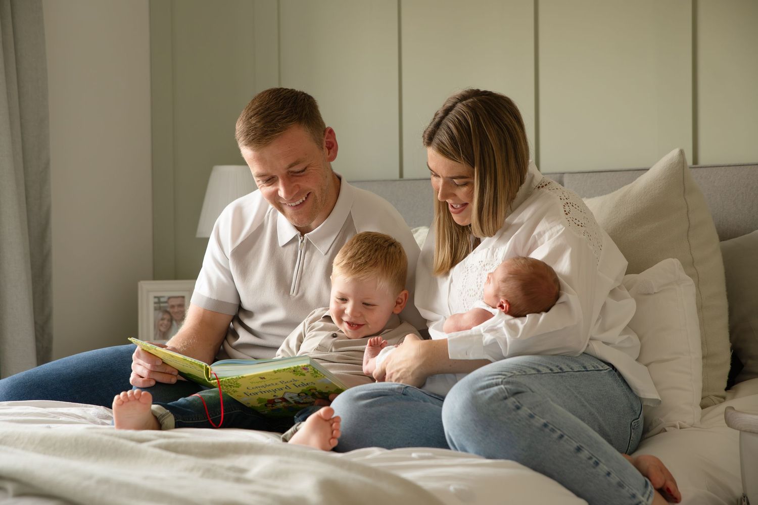Parent and child lying in bed reading a storybook together in a cozy white bedroom.
