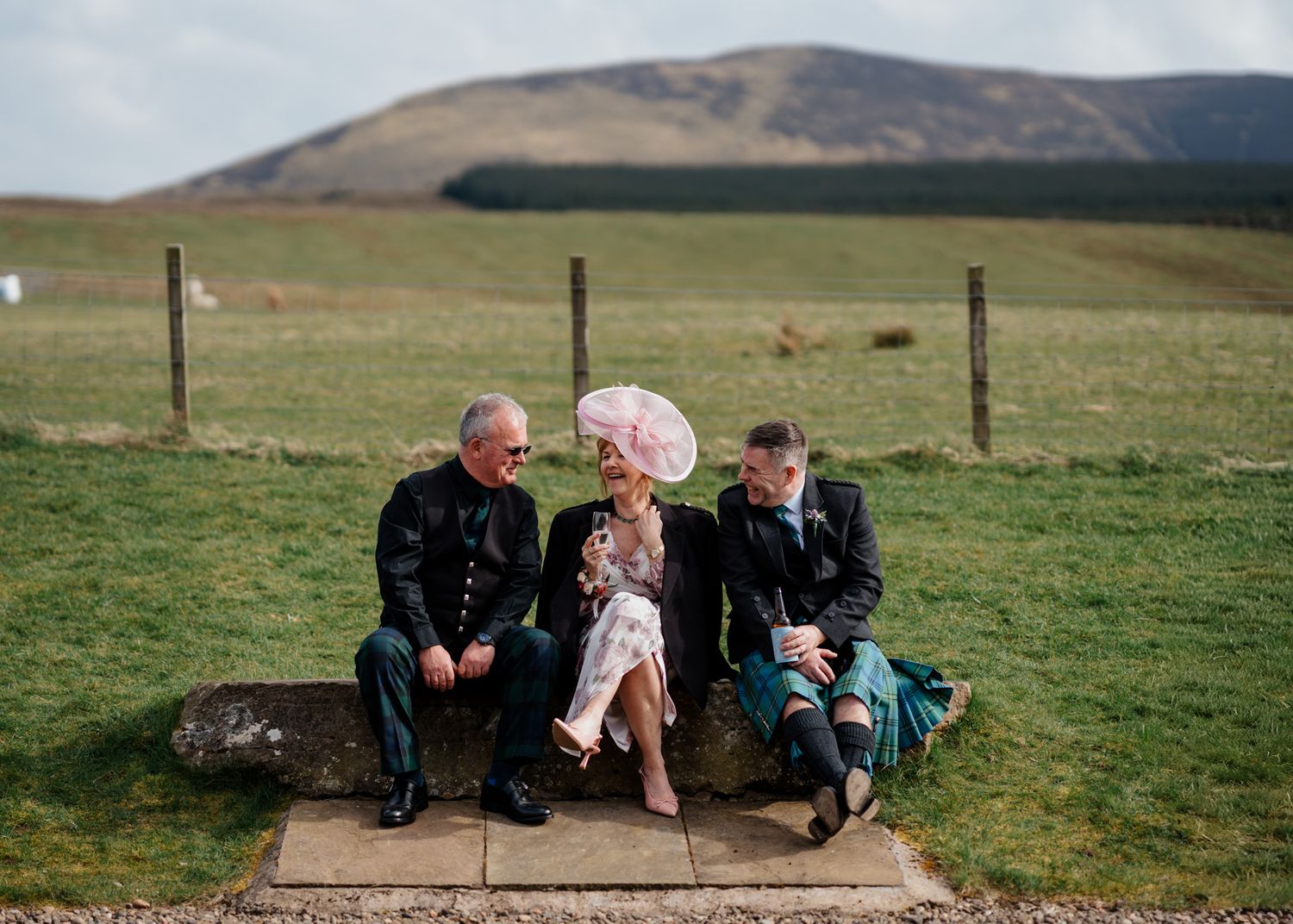 Three wedding guests sit on a stone bench in a Scottish Highland landscape, with rolling green hills in the background.