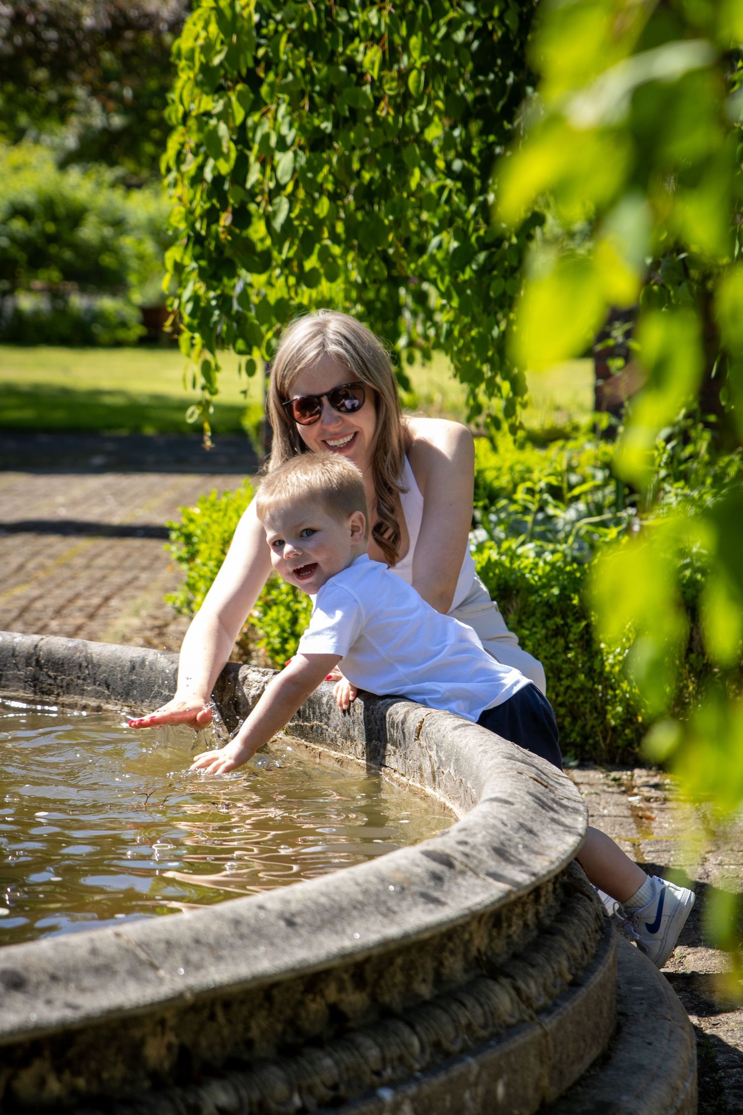 People playing and splashing at a stone water fountain in a sunny garden setting.