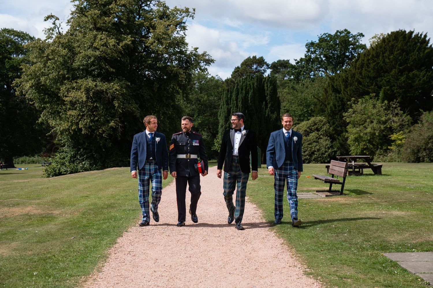 Four men in blue suits and tartan kilts walking along a garden path on a sunny day.