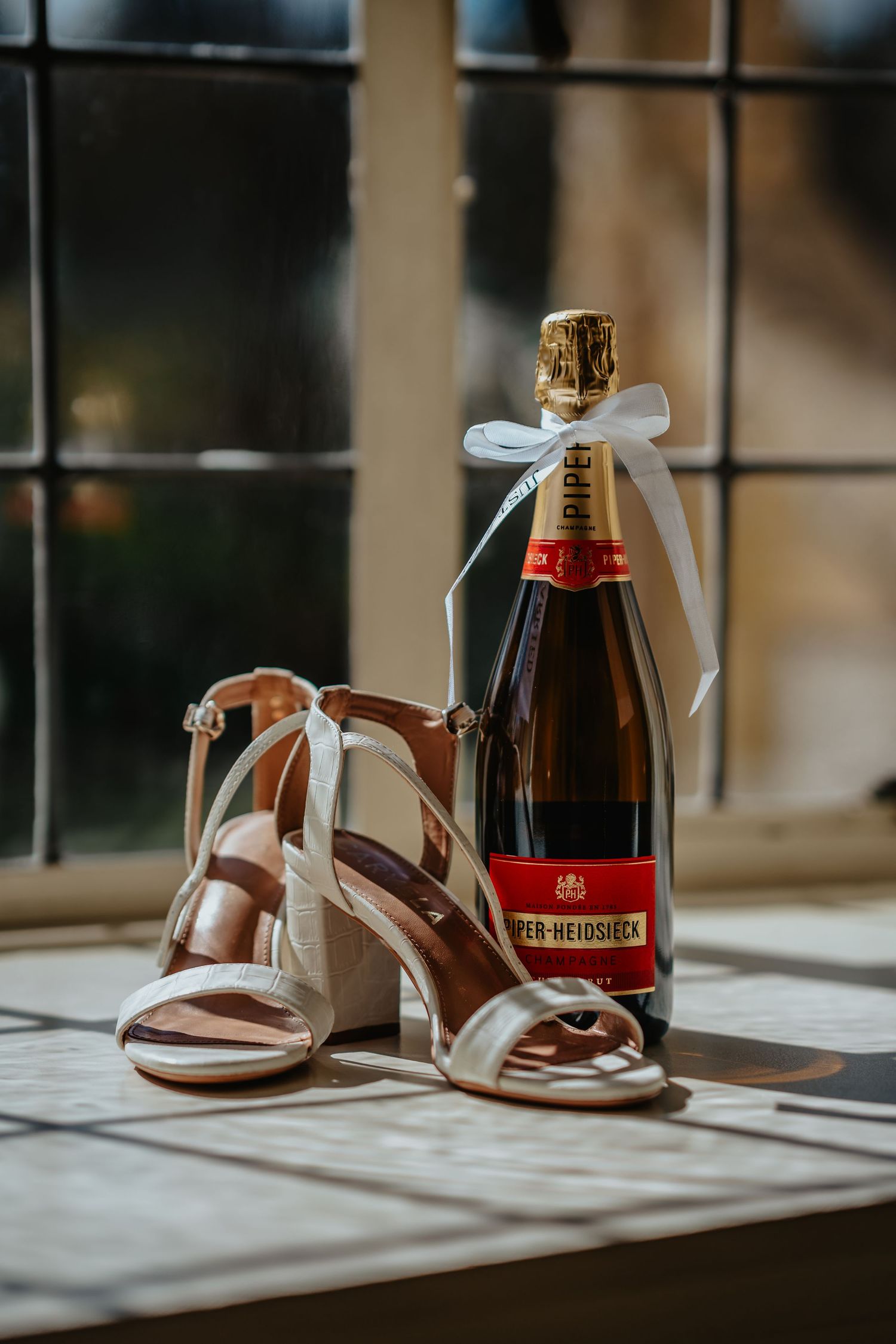 Elegant bridal heels beside a Piper-Heidsieck champagne bottle tied with white ribbon on a sunlit windowsill.