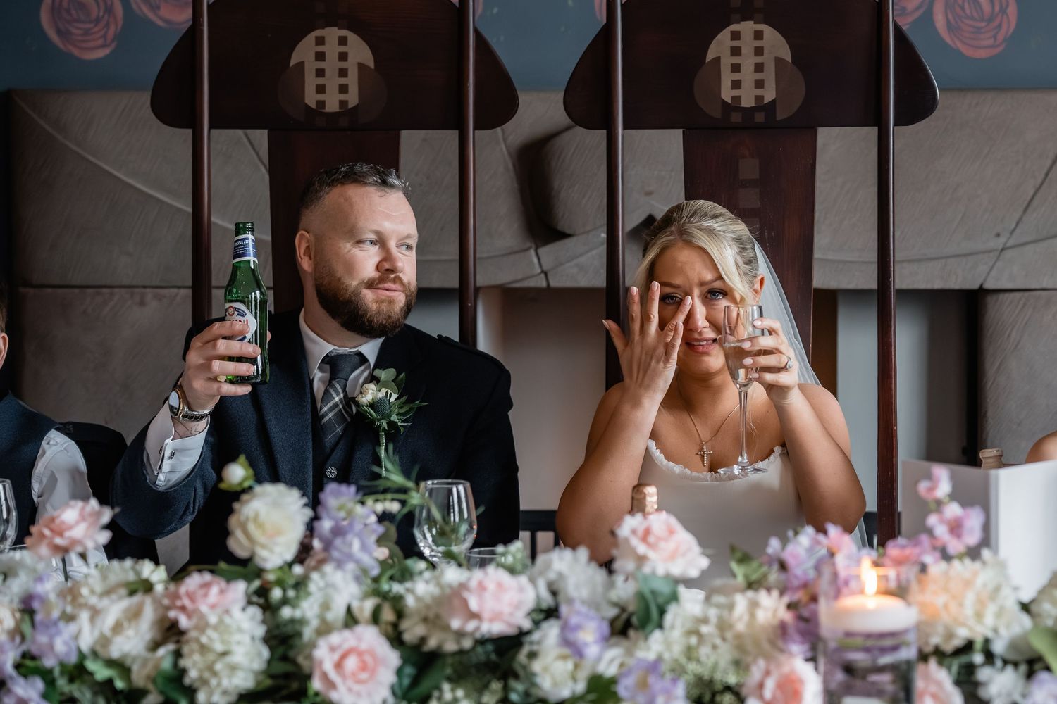 Emotional bride wiping tears beside groom holding beer bottle at wedding reception table with floral arrangements.