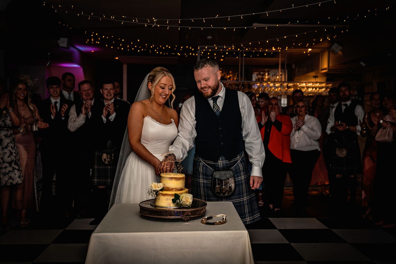 Bride and groom cutting wedding cake on checkered floor surrounded by guests at a Scottish wedding reception.
