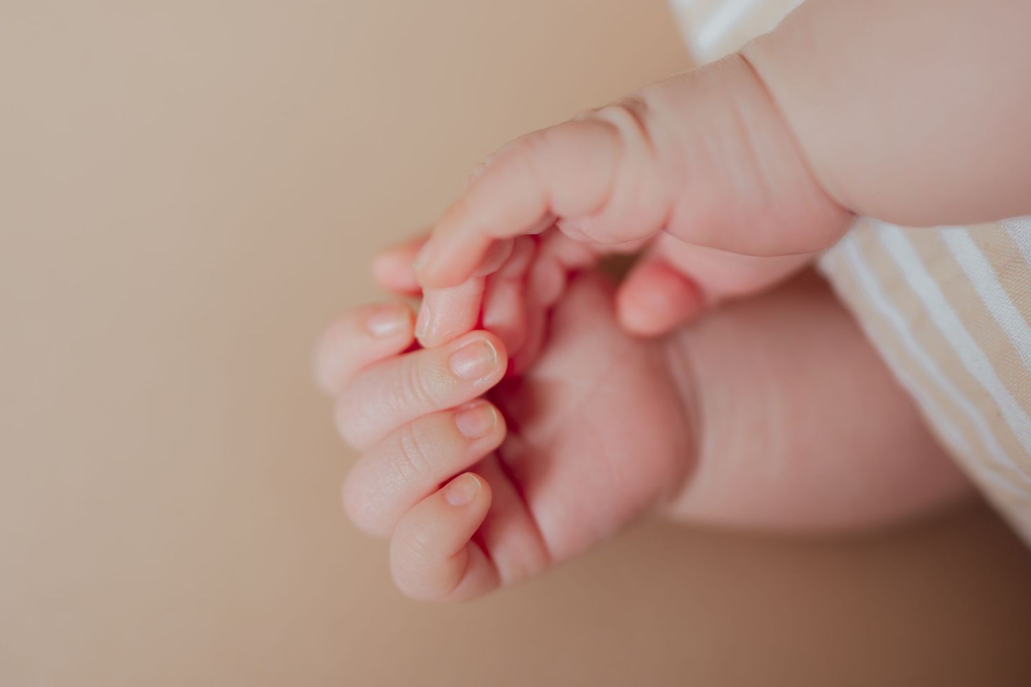 Close-up of a newborn baby's tiny delicate hands with soft pink fingers on a warm beige background.