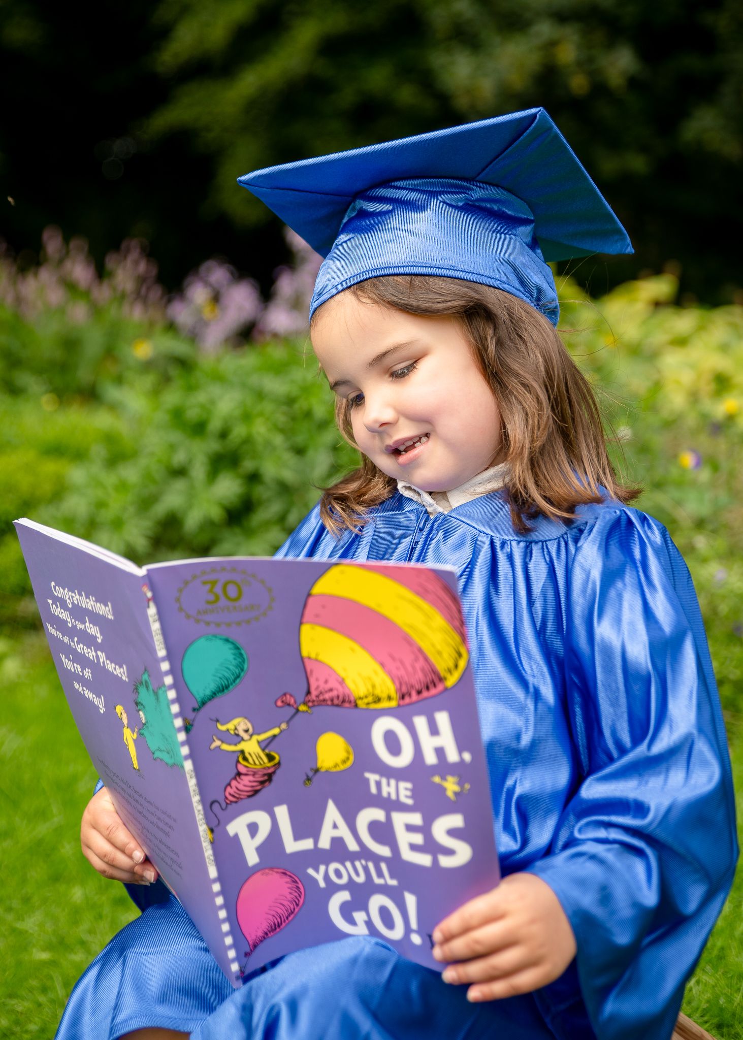 Young girl in blue graduation cap and gown smiling while reading Dr. Seuss Oh The Places Youll Go book outdoors.
