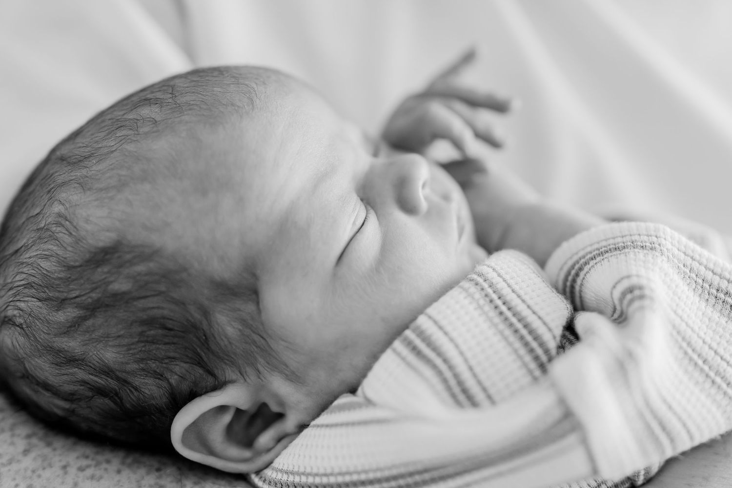 Black and white photo of a peaceful sleeping newborn baby with tiny hands raised, wearing a striped onesie.