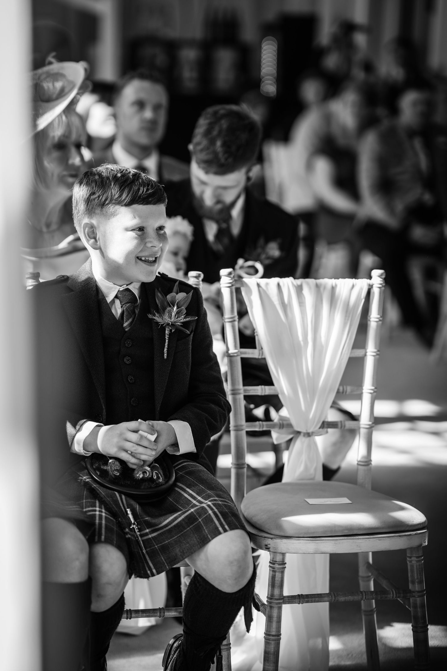 A smiling boy in a kilt and suit with boutonniere sits at a wedding ceremony in black and white photo.