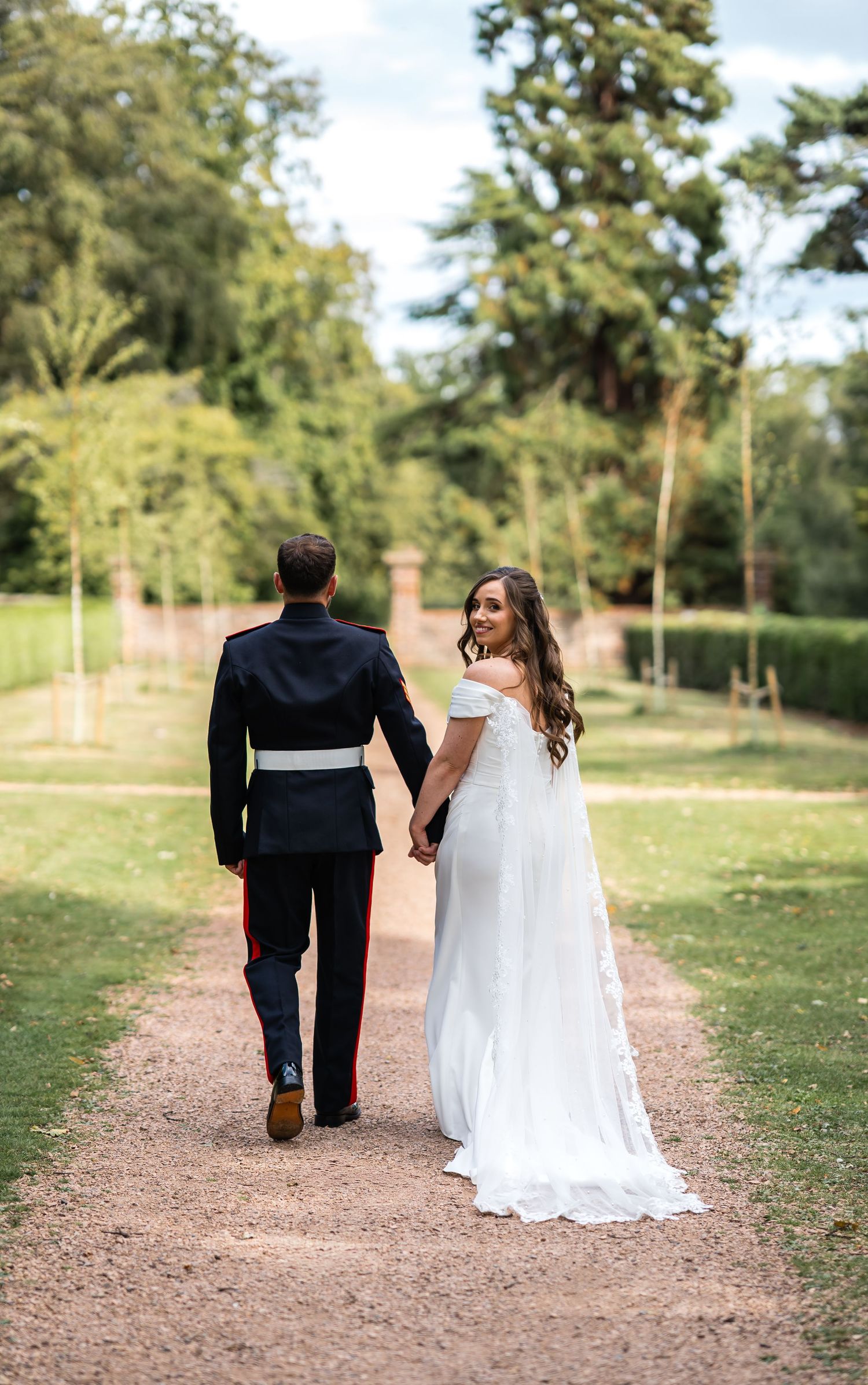 A bride in a white gown and groom in military uniform walk hand in hand along a tree-lined garden path.