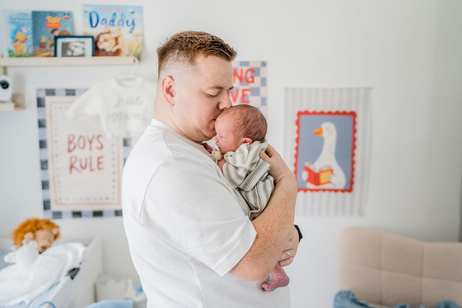 Father kissing newborn baby in nursery room with Boys Rule sign on wall.